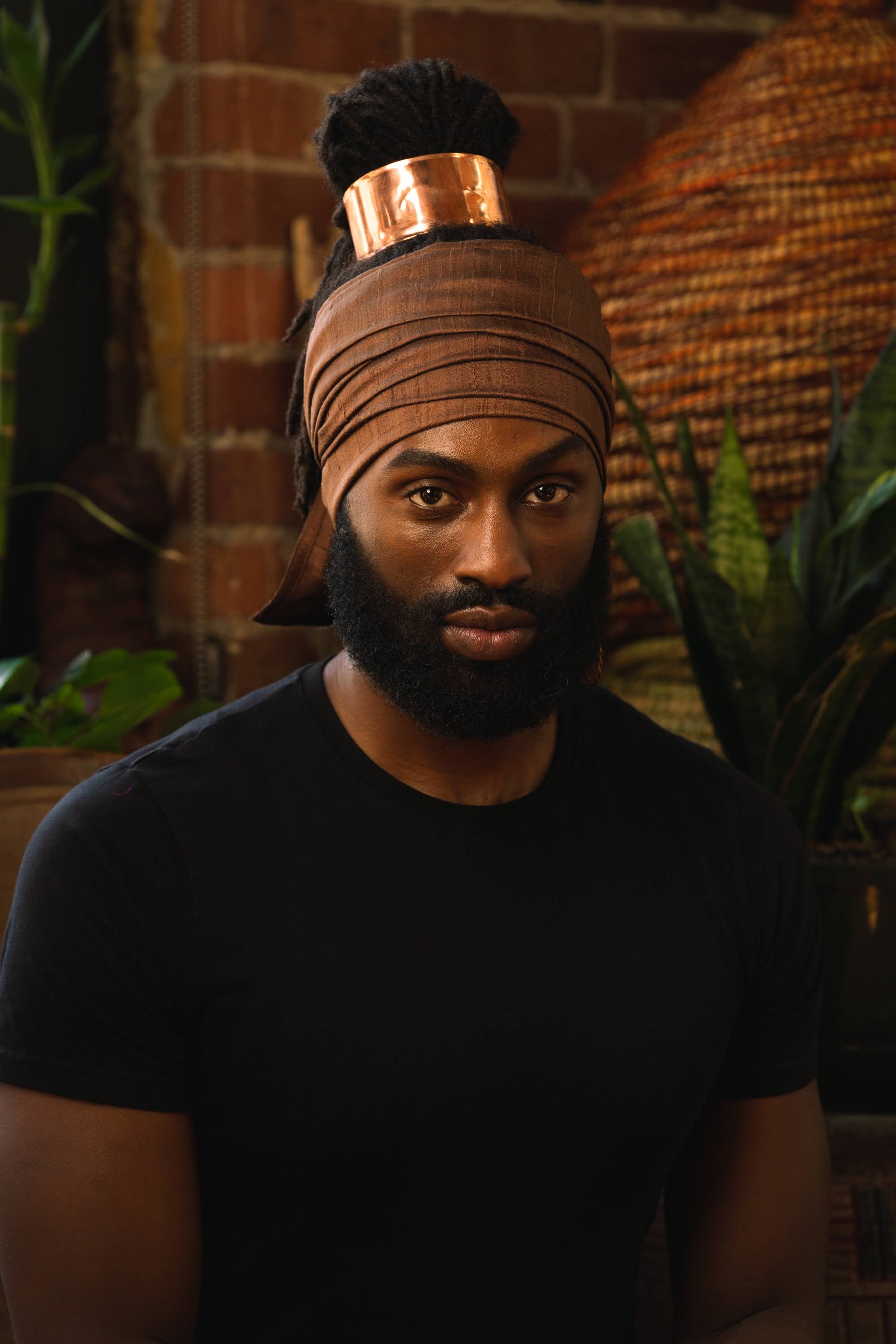 Bearded black man seated wearing brown silk head wrap with copper hair cuff around locs