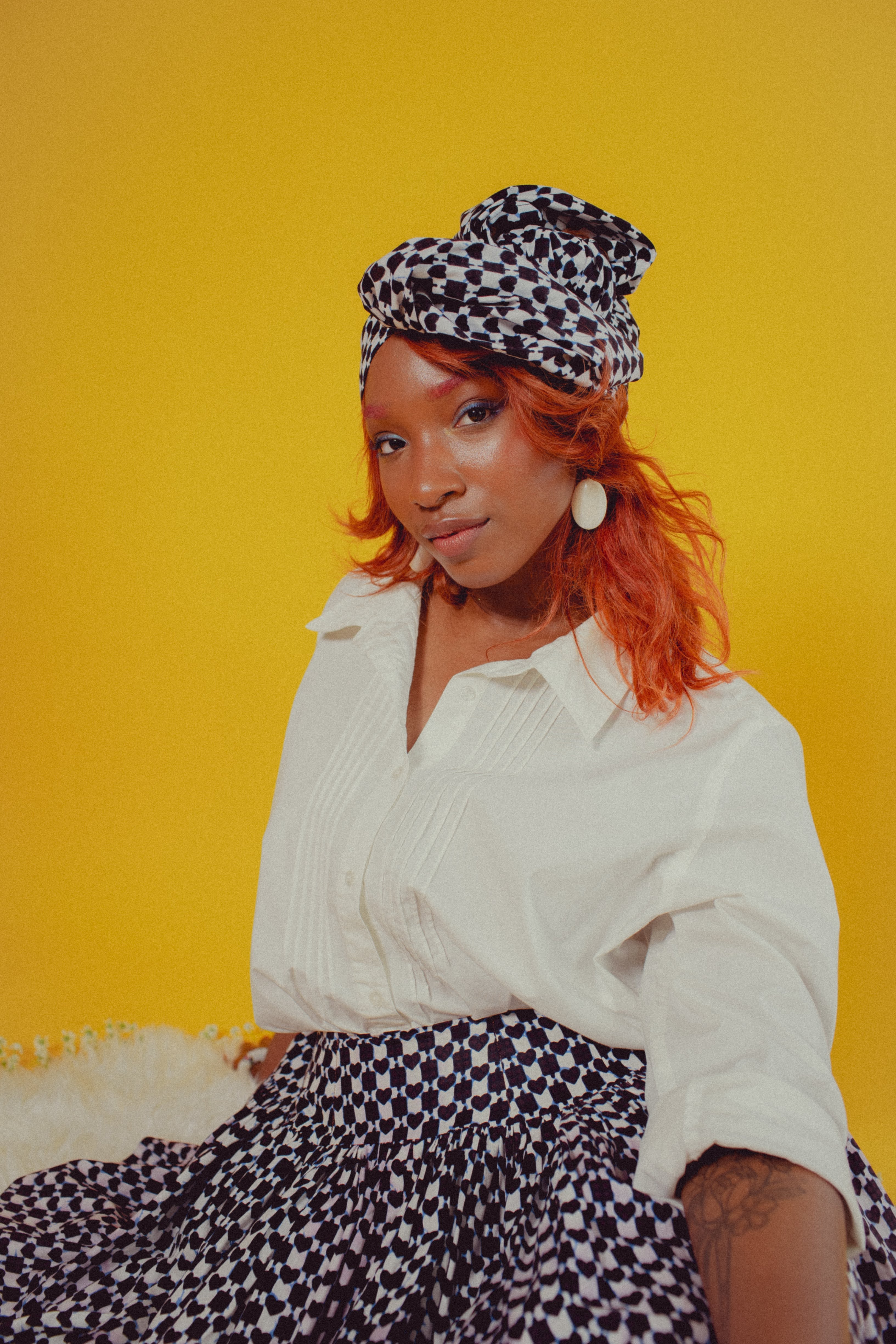 Black woman with red hair seated on fuzzy white rug wearing checked head wrap and matching checkered baby doll skirt with blue tulle