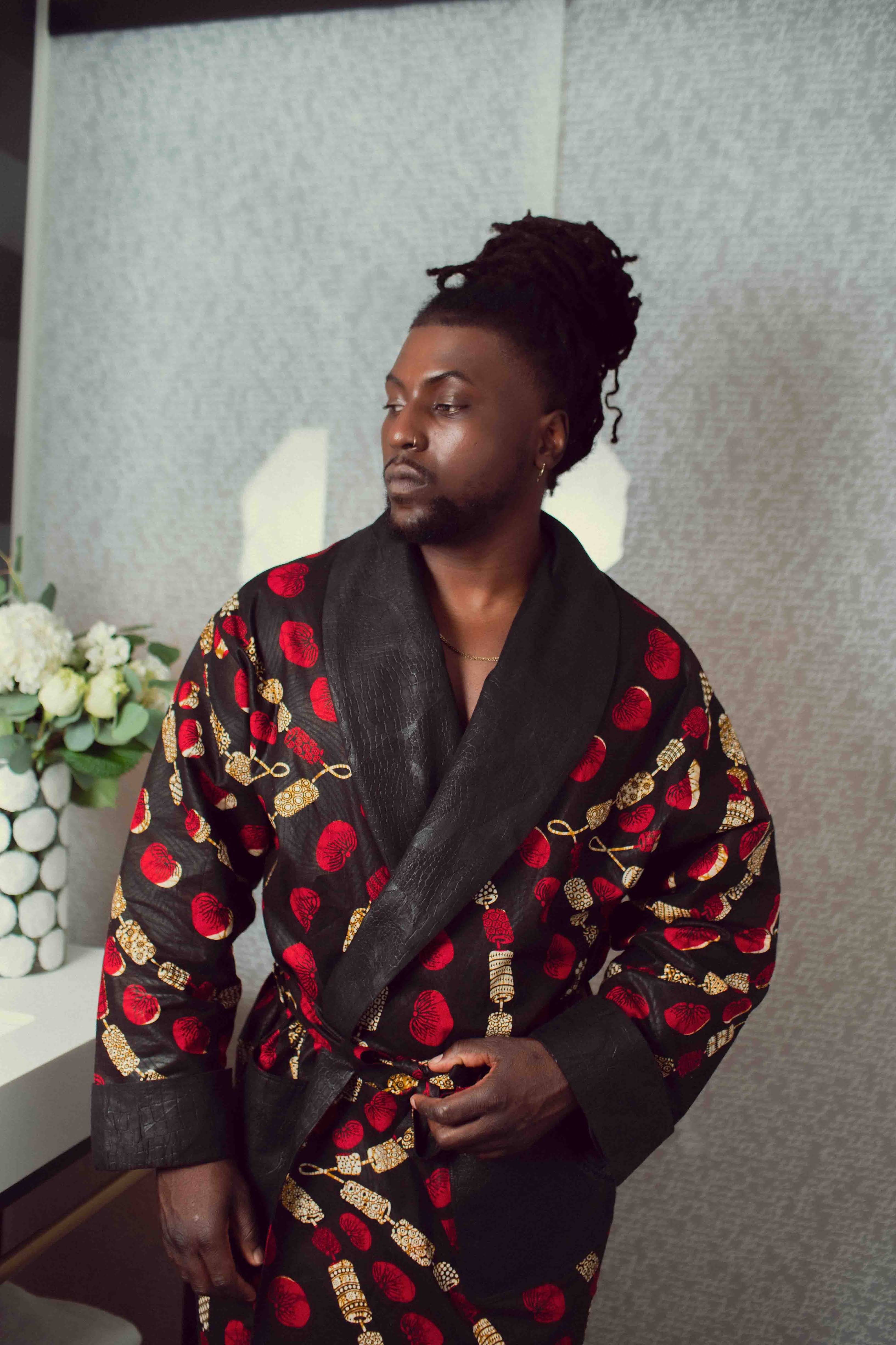 Black man with locs in a top bun wears small patterned red and cream smoke jacket with textured black cotton while in the bathroom